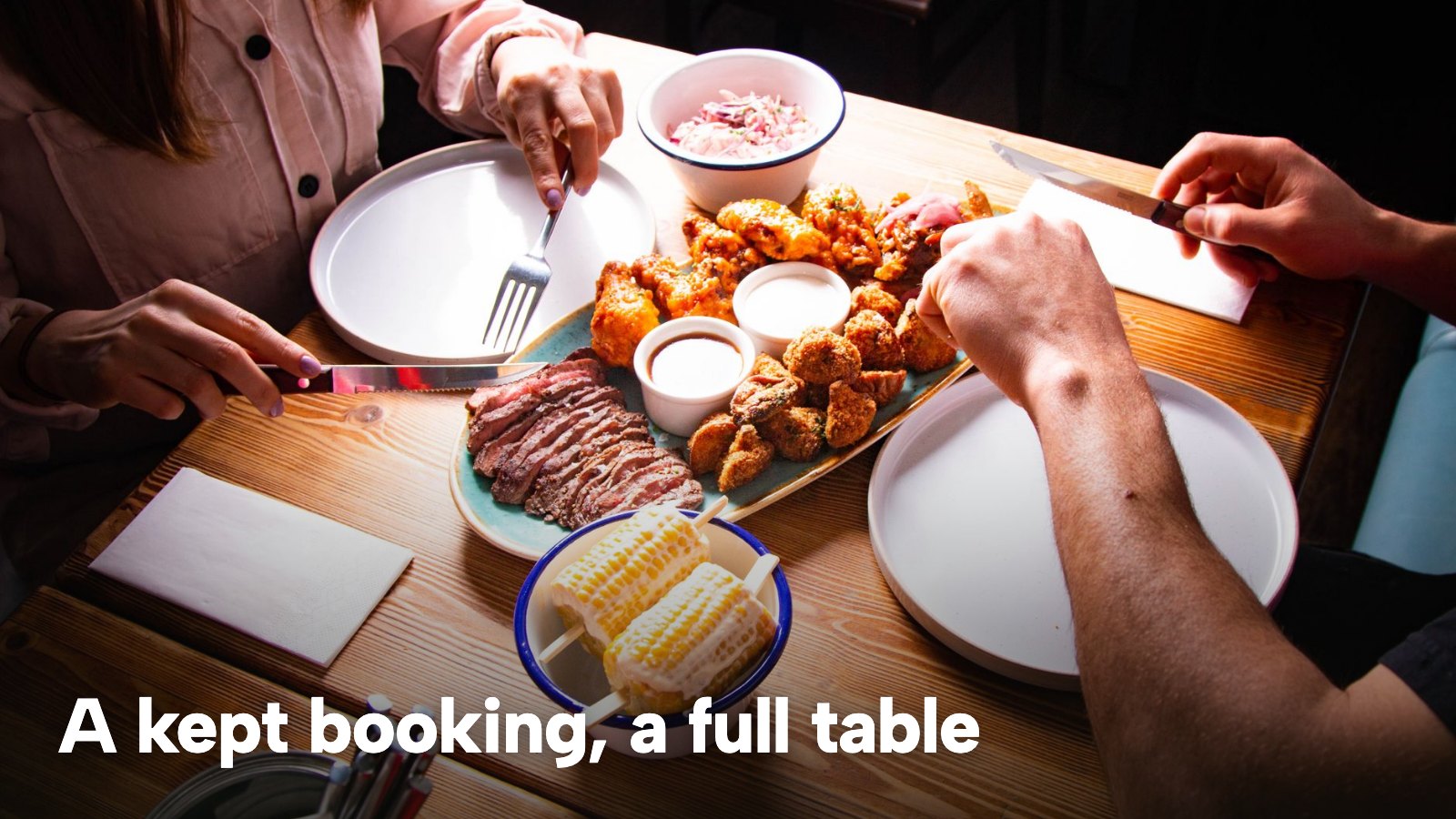 Two diners sharing steak and side dishes at a wooden restaurant table. The image carries the words "A kept booking, a full table".