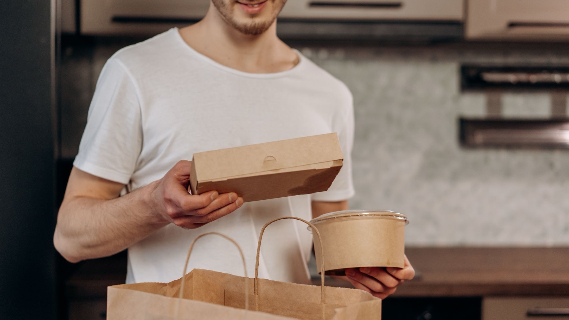 A man in a white shirt packing takeaway food containers in a kitchen.