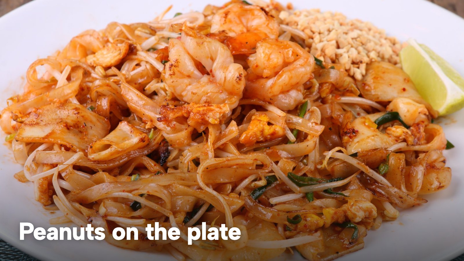 A plate of shrimp pad thai topped with crushed peanuts and a lime wedge. The image carries the words "Peanuts on the plate".